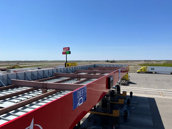 American long-nose 53-foot conventional cab semi-truck parked at a truck stop with an empty highway stretching into the distance