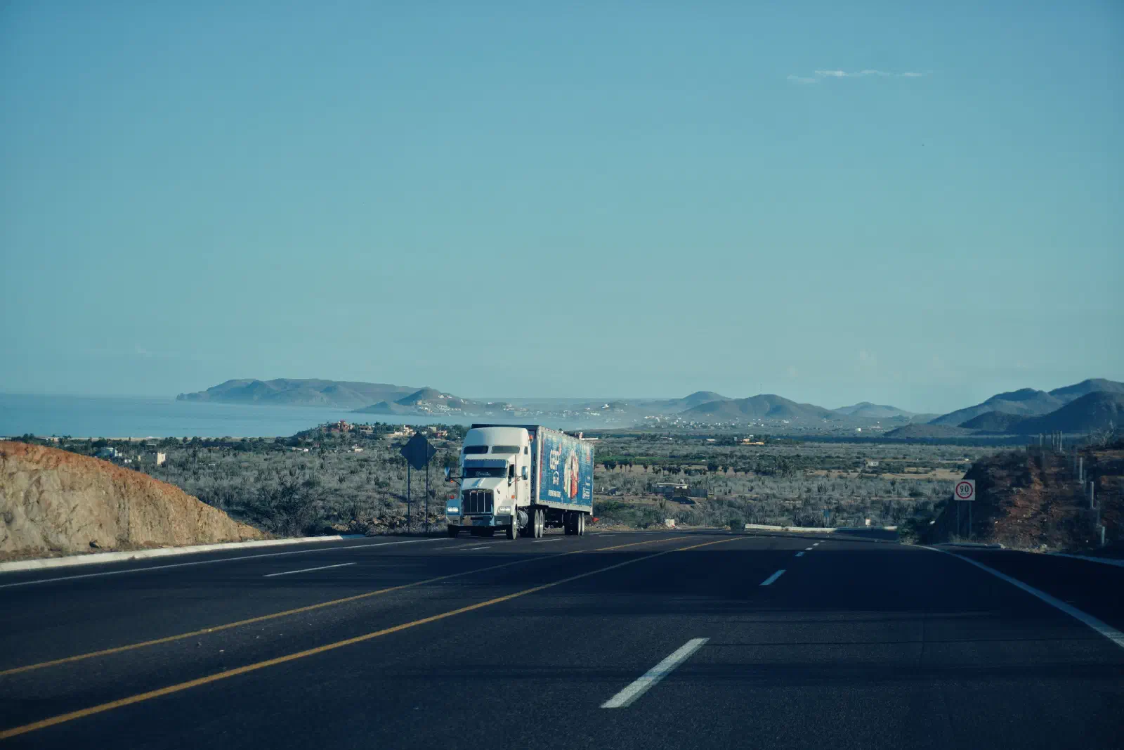 American long-nose 53-foot conventional cab semi-truck parked at a truck stop with an empty highway stretching into the distance